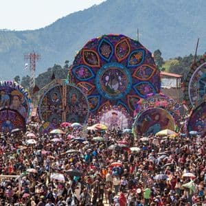 Eine riesige Menschenmenge versammelt sich auf einem Open-Air-Festival, um mehrere riesige, bunte, verzierte Drachen zu bestaunen, die vor einer Bergkette ausgestellt sind.