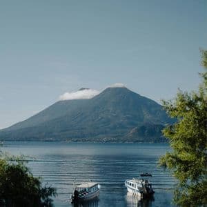 Zwei Motorboote treiben auf einem ruhigen See vor einem großen Berg mit wolkenbedecktem Gipfel, umrahmt von grünem Laub.