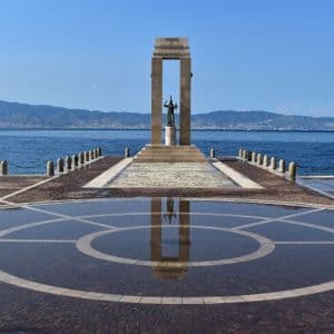 Un monumento su un molo di pietra si riflette in una grande pozzanghera, con il mare e le montagne sullo sfondo.