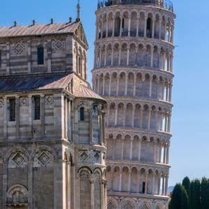 An einem sonnigen Tag steht ein schiefer, weißer Marmorglockenturm neben einer kunstvollen Steinkathedrale, auf dessen Rasen sich Menschen versammelt haben.