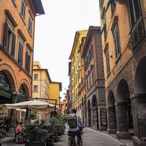 Ein Mann mit Rucksack fährt mit dem Fahrrad eine enge Straße entlang, die von hohen, farbenfrohen Gebäuden mit Bogengängen gesäumt ist.