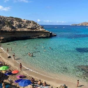 Gente tomando el sol en una playa de arena y nadando en las claras aguas turquesas de una cala rocosa en un día soleado.