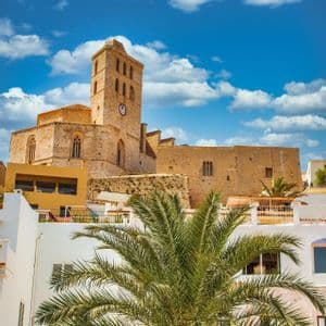 Una catedral de piedra con torre del reloj en una colina, vista sobre edificios blancos y una palmera bajo un cielo azul nublado.