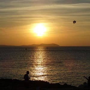 Siluetas de personas en una costa rocosa observan un atardecer dorado sobre el mar, con un parapente visible en el cielo.