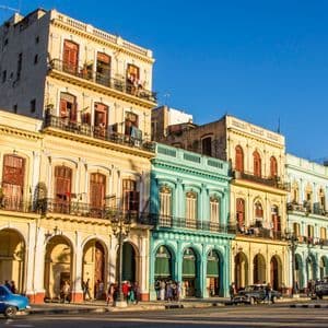 Des bâtiments colorés de style colonial bordent une rue de la ville où des gens se promènent et des voitures anciennes circulent sous un ciel bleu clair.