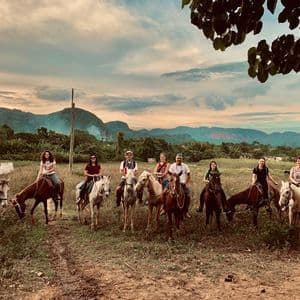 Un grupo de viaje de WeRoad posa a caballo en un campo de hierba con montañas verdes de fondo bajo un cielo nublado.
