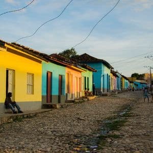 Una calle empedrada bordeada de casas coloridas en verde, amarillo y azul, con gente relajándose al aire libre durante el atardecer.