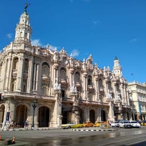 An ornate, light-colored classical building with towers stands on a city street corner. Vintage yellow taxis and other cars are parked in front under a blue sky.