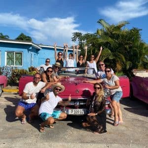 Un gruppo WeRoad in viaggio sorride e posa per una foto intorno a una decappottabile rosa d'epoca, davanti a un edificio blu.
