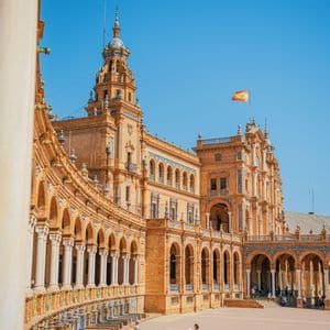 Ein imposantes orangefarbenes Backsteingebäude mit Bögen und einer spanischen Flagge auf dem Dach, von einem sonnigen Platz mit ein paar Menschen aus gesehen.