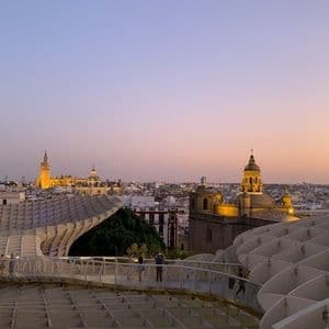Menschen gehen auf einem modernen Hochweg mit Blick auf eine Stadt-Skyline mit beleuchteten Türmen bei Sonnenuntergang.