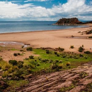 A wide, sandy beach beside a calm body of water, with green grassy patches and a rocky headland under a partly cloudy sky.