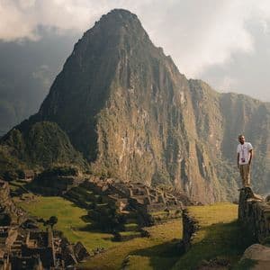 A man in a white t-shirt stands on a stone wall overlooking ancient terraced ruins and a steep mountain peak under a cloudy sky.