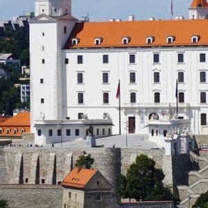 Un gran castillo blanco con techos de tejas naranjas y torres en las esquinas se asienta sobre una colina de piedra fortificada bajo un cielo parcialmente nublado.