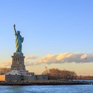 Die Freiheitsstatue steht auf ihrem Sockel auf Liberty Island, im Licht des Sonnenuntergangs vor blauem Himmel mit Wolken.