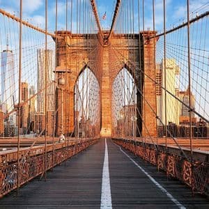 An empty wooden walkway on a suspension bridge with stone arches leading towards a city skyline at sunrise.