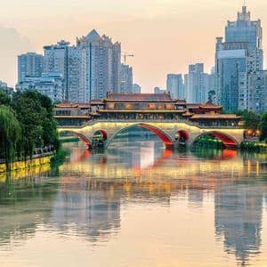 An illuminated traditional bridge with red arches spans a river, reflecting on the water with a modern city skyline in the background.