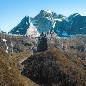 Jagged, snow-capped mountains tower over a valley with autumn-colored forests under a clear blue sky.