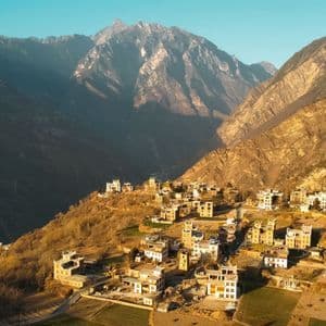 A village of traditional, multi-story buildings clings to a sunlit hillside at the base of a rugged mountain range.