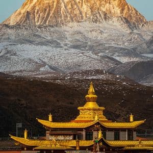 A golden temple sits in the foreground with a massive, snow-dusted mountain range illuminated by sunlight in the background.