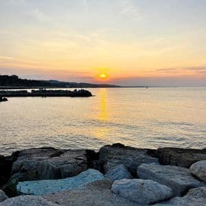 Il sole tramonta su una distesa d'acqua calma, osservato da un frangiflutti roccioso, proiettando un riflesso dorato sulla superficie.