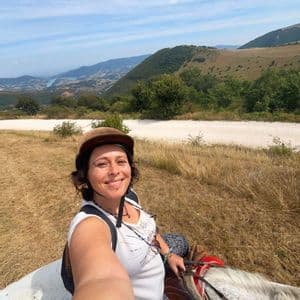 Una donna sorridente con un casco si scatta un selfie mentre cavalca un cavallo bianco su una collina, con una valle e un lago sullo sfondo.