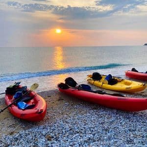 Kayak rossi e gialli riposano su una spiaggia di ciottoli con onde dolci mentre il sole tramonta sull'oceano sotto un cielo nuvoloso.