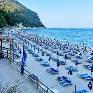 File di ombrelloni e lettini da spiaggia blu e bianchi costeggiano una spiaggia sabbiosa vicino al mare, con una collina boscosa sullo sfondo.