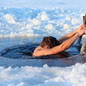 Eine Person hält sich an einer Holzleiter fest und taucht in ein Eisloch in einem zugefrorenen, schneebedeckten Gewässer ein.