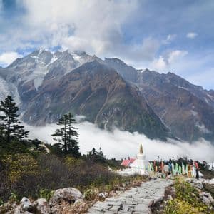 A stone path leads to a white stupa and prayer flags, with snow-capped mountains and a glacier in the background.