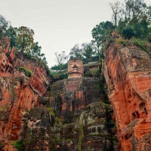 A giant Buddha statue carved into a red sandstone cliff, nestled between two rock faces with trees on top.