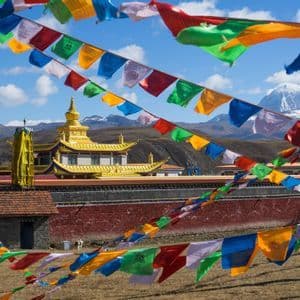 Colorful prayer flags blow in the wind, framing a view of a golden-roofed monastery and distant snow-capped mountains.
