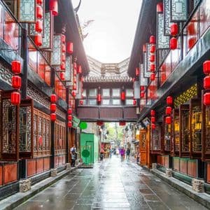 A narrow street with traditional architecture is lined on both sides with hanging red lanterns, reflecting on the wet pavement.