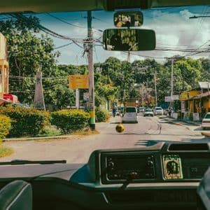 Vista dal cruscotto di un'auto su una strada di città con auto, edifici colorati e alberi verdi rigogliosi.