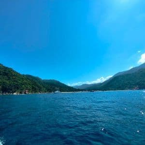Uno scorcio dal mare di una costa montuosa e alberata, sotto un cielo azzurro e soleggiato, con una piccola barca in lontananza.