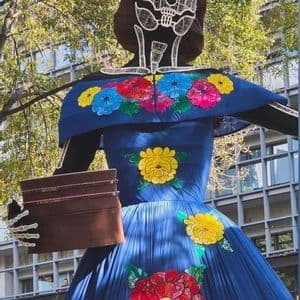 Una gran figura de Calavera Catrina con un rostro de calavera blanco y un vestido azul adornado con flores coloridas se encuentra al aire libre.