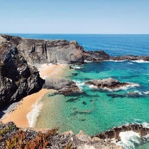 Veduta aerea di una cala sabbiosa e appartata, con acque cristalline color turchese, circondata da scogliere rocciose sotto un cielo azzurro.