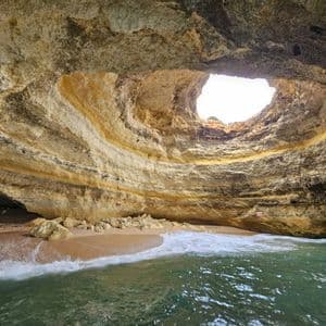 Le onde accarezzano una piccola spiaggia sabbiosa all'interno di una grande grotta marina, dotata di un'apertura rotonda verso il cielo nel suo soffitto.