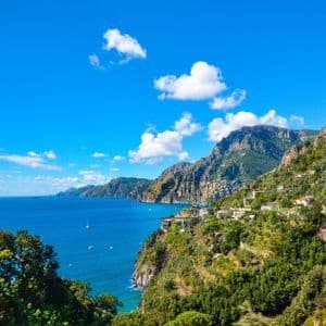 Una costa verde e montuosa, costellata di edifici bianchi, digrada verso un mare azzurro calmo sotto un cielo luminoso con poche nuvole.