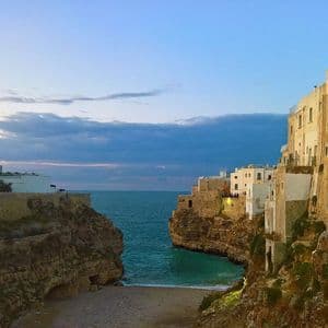 Edifici bianchi costruiti su scogliere rocciose circondano una piccola baia e una spiaggia sabbiosa che si affacciano sul mare aperto al crepuscolo.