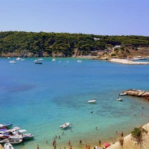 Una veduta dall'alto di una baia costiera dove le persone nuotano in acque turchesi, con barche ormeggiate a un molo e ancorate nel blu profondo del mare.