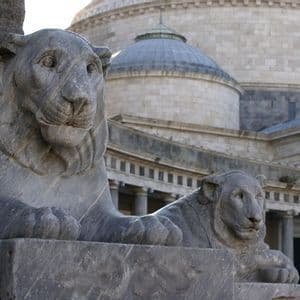 Due statue di leoni in pietra grigia su piedistalli davanti a un edificio classico con una cupola.