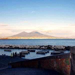 Una vista su una baia verso un grande vulcano, con una città ai suoi piedi e piccole imbarcazioni ormeggiate in un porto in primo piano.