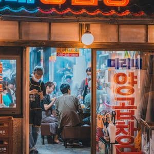 An open-front Korean restaurant bustling with customers at night, viewed from the street under glowing neon signs.