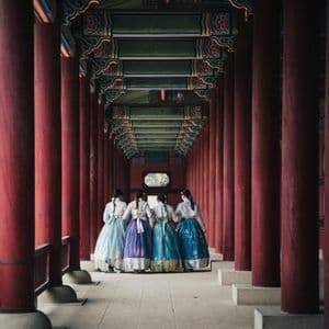 A WeRoad group trip of four women in colorful traditional hanbok dresses, seen from behind in a long corridor with red pillars.