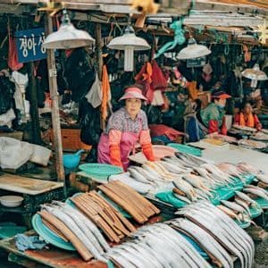 A female fish vendor in a pink apron and visor stands behind a stall filled with fresh fish at an outdoor market.