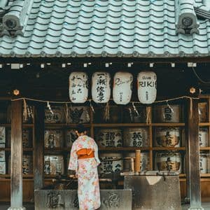 A woman in a floral kimono, seen from behind, stands at a water basin in front of a Japanese shrine with hanging lanterns.