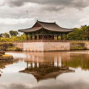 A traditional Korean pavilion on a stone foundation sits over a calm lake, its reflection visible in the water under a cloudy sky.