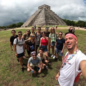 Un groupe WeRoad prend un selfie souriant dans un champ verdoyant, devant une grande pyramide antique en pierre, sous un ciel nuageux.