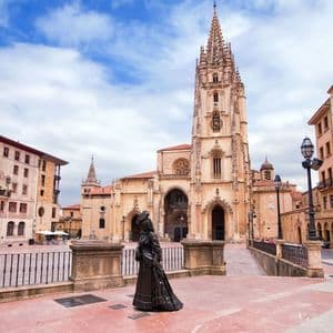 Una estatua de bronce de una mujer se alza en una plaza pavimentada frente a una ornamentada catedral gótica bajo un cielo parcialmente nublado.